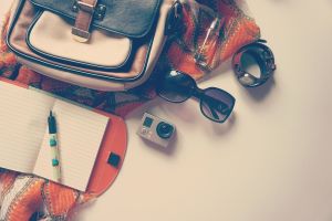 Widok z góry na leżący plecak, okulary, aparat, zegarek i notes z długopisem