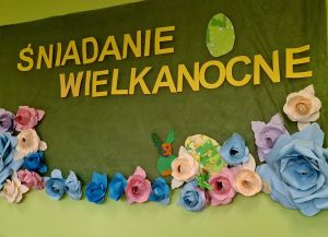 Napis na zielonym tle ŚNIADANIE WIELKANOCNE. Pod napisem znajdują się kwiaty, a nad napisem pokolorowane jajko.