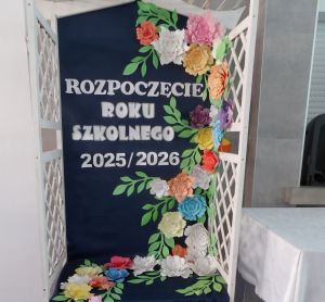 Ścianka dekoracyjna ozdobiona kwiatami. Na środku znajduje się napis: Rozpoczęcie roku szkolnego 2025/2026